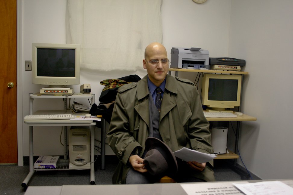 A man in a trench coat sits in an office holding a fedora and audition pages.
