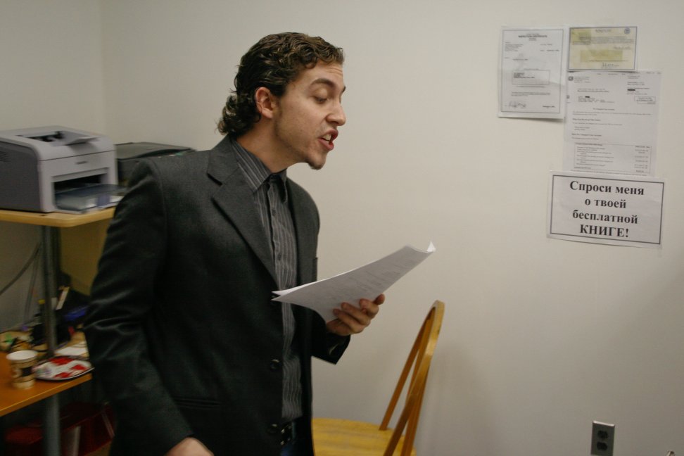 A man in a dark suit stands in an office reading from a script.