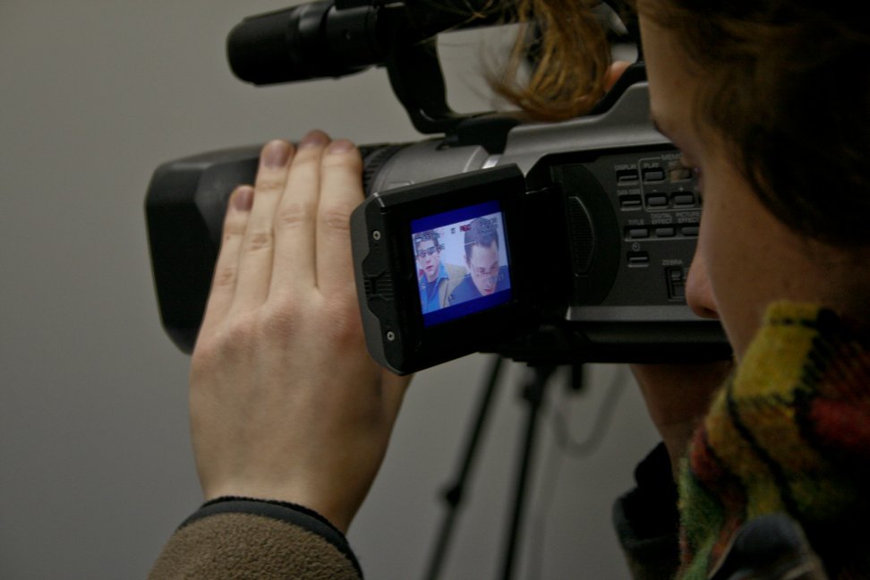 A camera operator films an audition, with the performer's face visible on the camcorder screen.