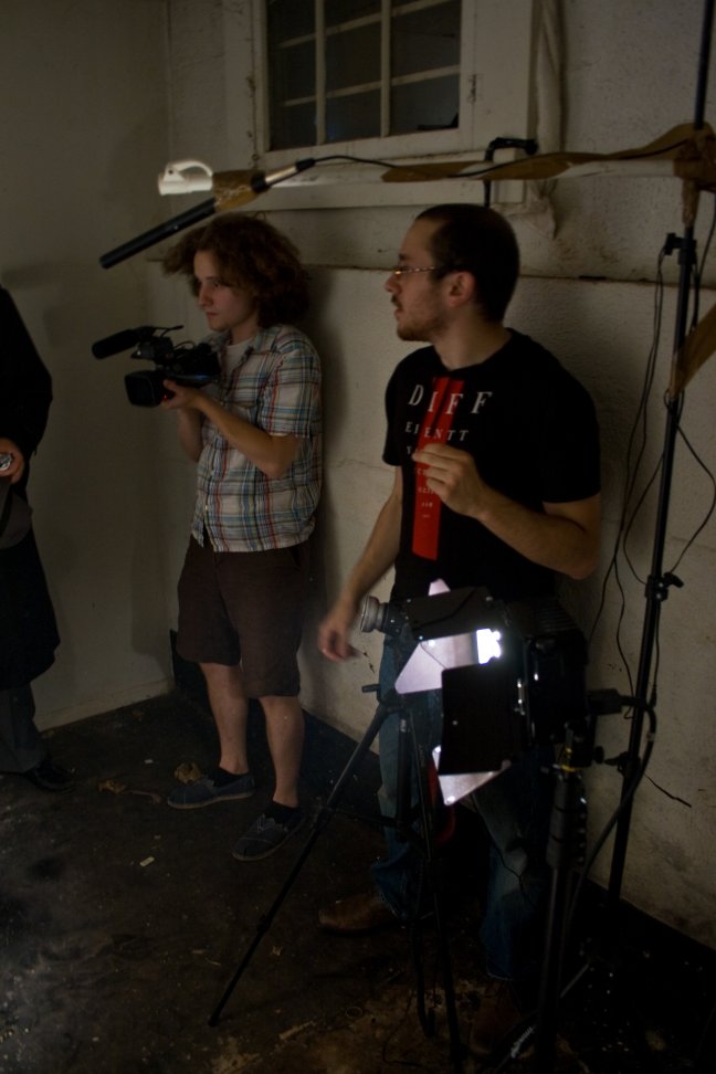 A small camera crew prepares lights and equipment in a dark industrial room.