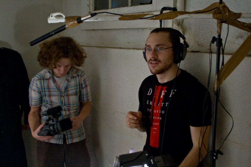 Two crew members stand beneath a boom microphone, one holding a camera and the other wearing headphones.