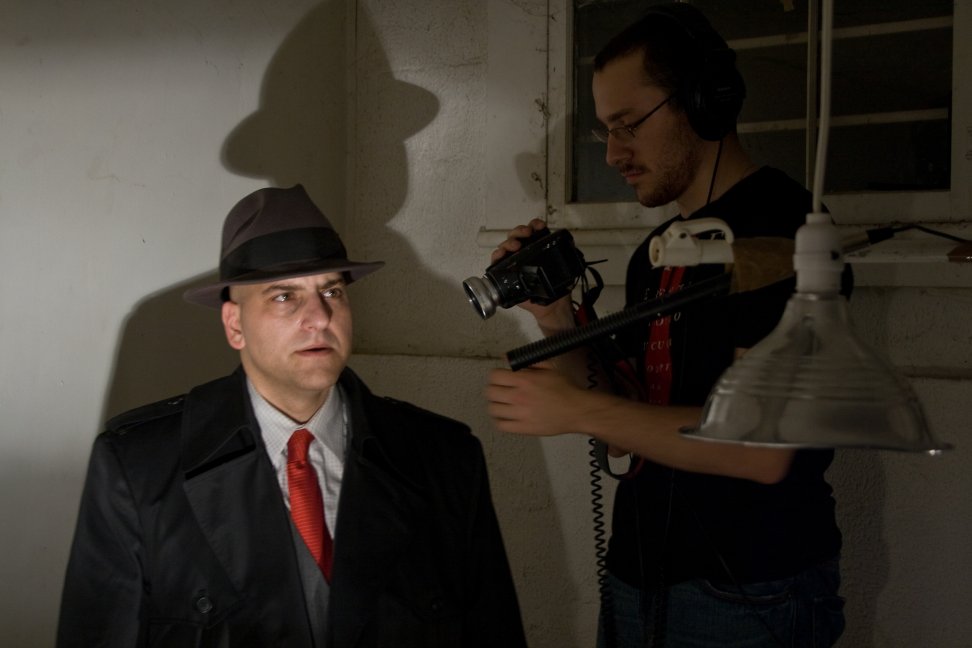 A camera operator works beside a performer in a fedora, trench coat, and red tie beneath a boom microphone.