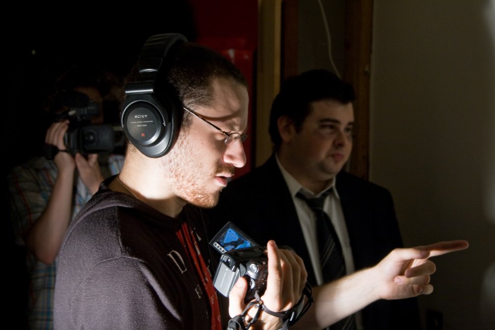 A crew member wearing headphones points while holding a camera, with a suited performer behind him.