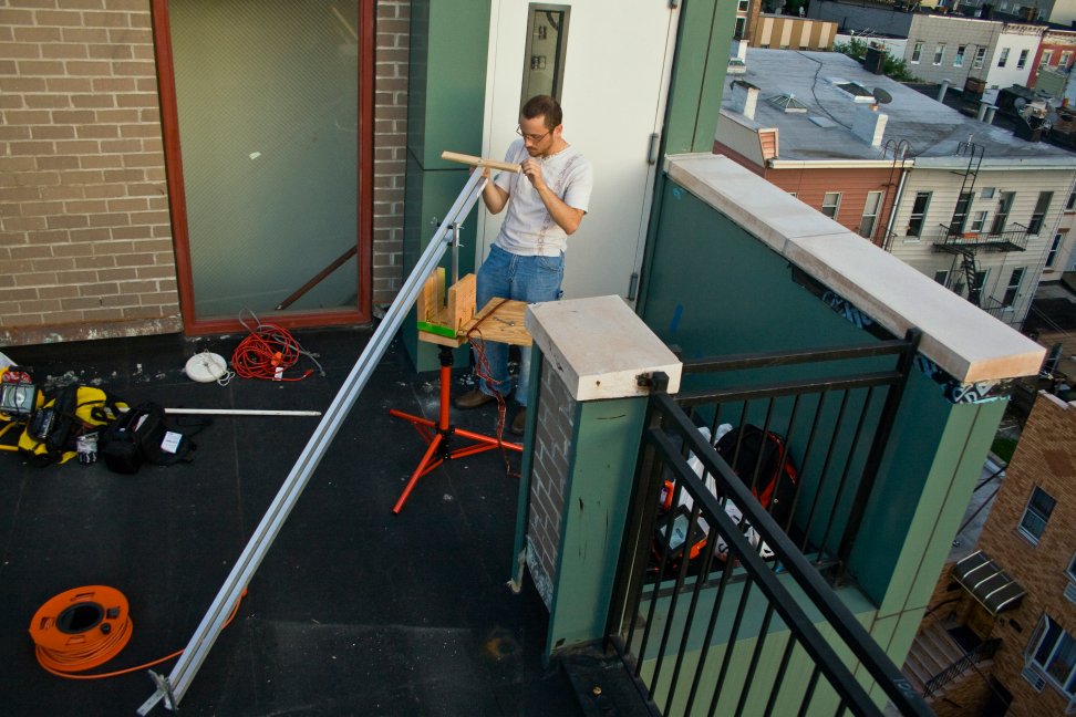 A crew member assembles a metal camera rig on a balcony above the city.