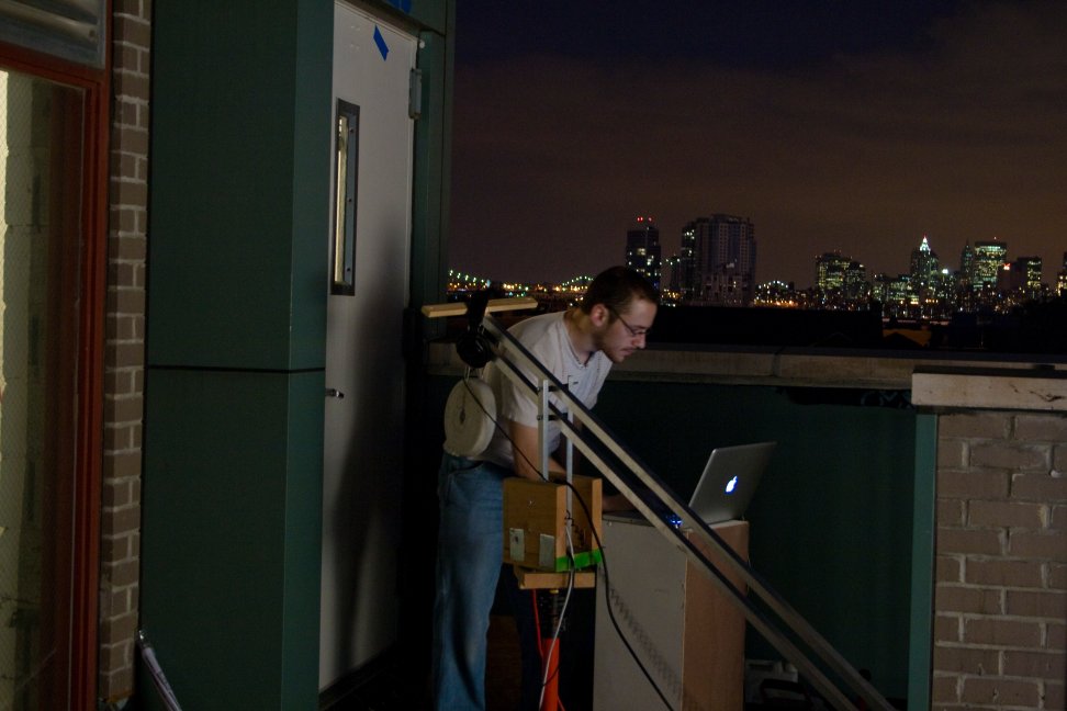 A crew member works beside a camera rig and laptop on a balcony overlooking the nighttime skyline.