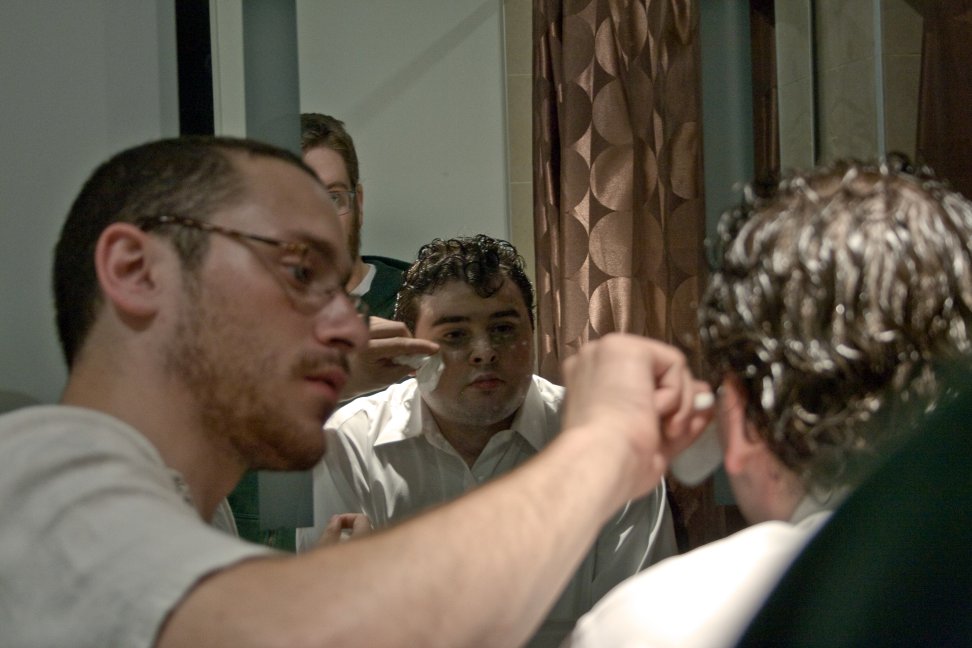 A makeup artist works on a seated performer in front of a bathroom mirror.