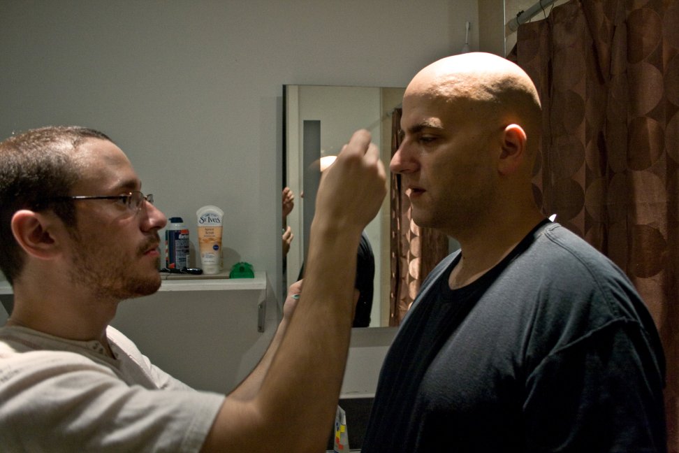 A makeup artist applies makeup to a bald performer before a scene.