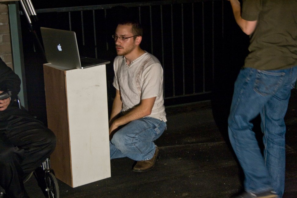 A crouching crew member watches a laptop mounted on a tall stand during the rooftop shoot.