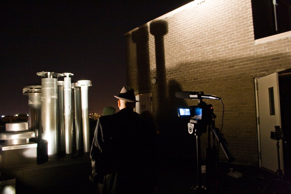 A camera and silhouetted performers stand beneath dramatic shadows on a nighttime rooftop.