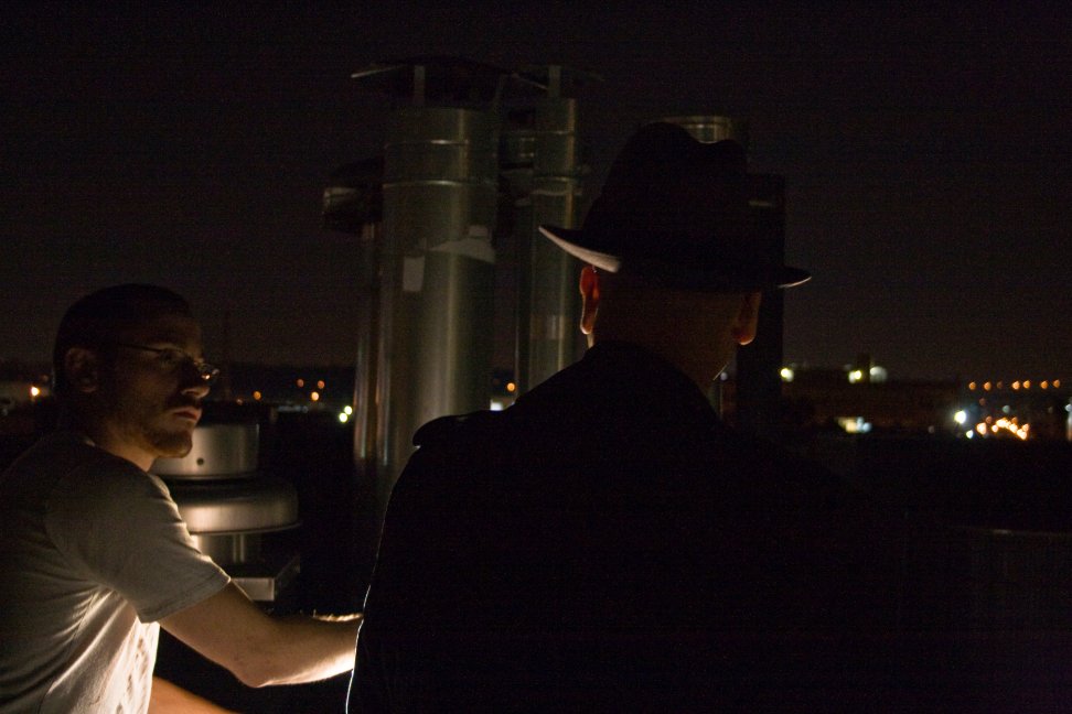A crew member guides a performer in a fedora and trench coat during a rooftop scene.