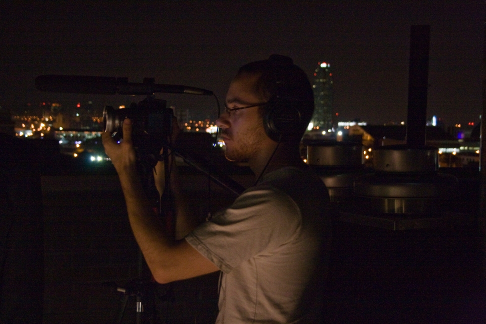 A camera operator records footage on a rooftop against distant city lights.