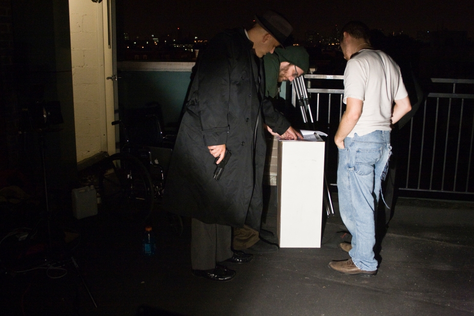 A costumed performer and two crew members inspect a rooftop setup beside a tall white equipment stand.