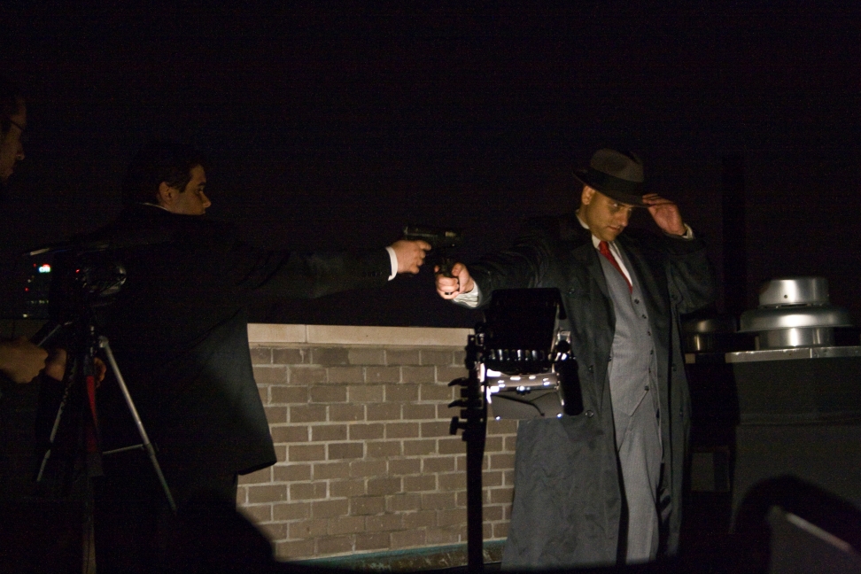 Two performers aim prop pistols during a nighttime rooftop scene as a camera records them.