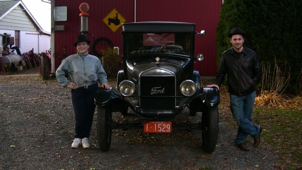 Two men in period-style hats pose beside a vintage Ford automobile outside a red barn.