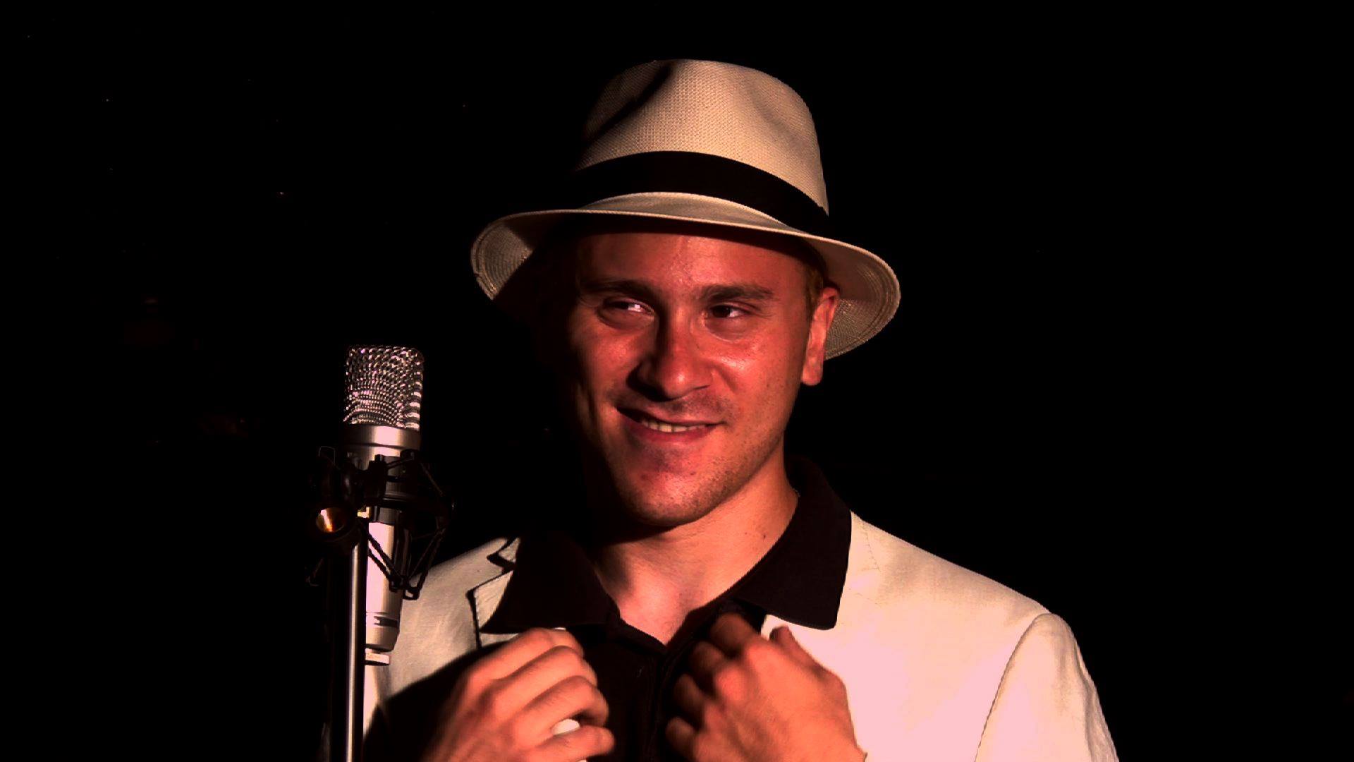 A smiling performer in a white fedora poses at a vintage microphone against a black background.