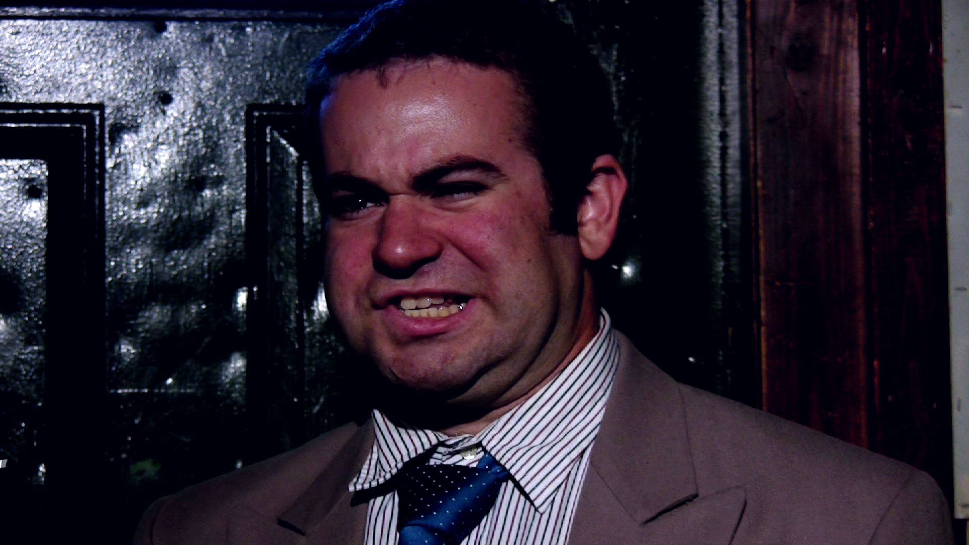 A man in a suit and blue tie grimaces in a stylized dark portrait.