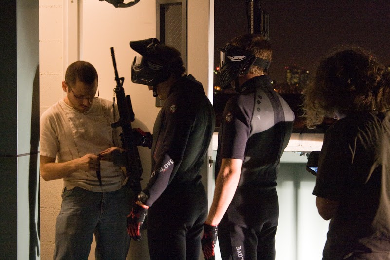A crew member adjusts a prop rifle while two performers in black costumes prepare for a scene.