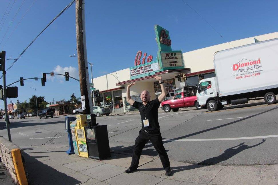 A man wearing an event badge poses with his arms raised outside a neighborhood cinema.