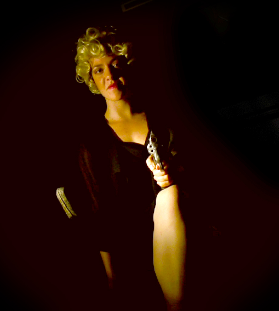 Blonde woman in a dark vintage costume holds a prop pistol in deep shadow.